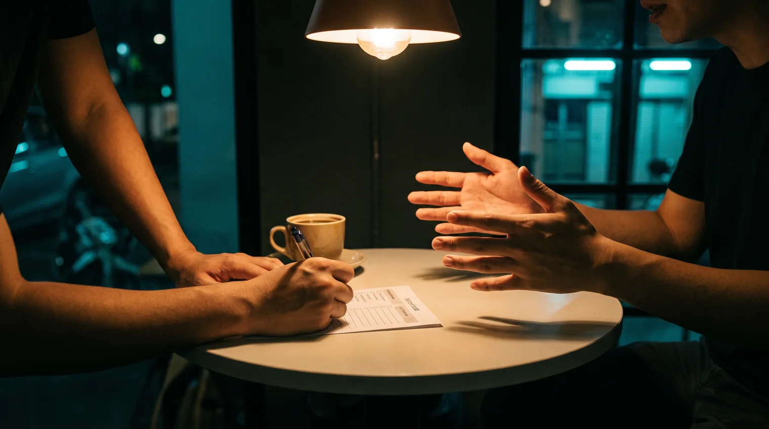 Two people at a cafe table at night, one writing on a printed checklist while the other explains