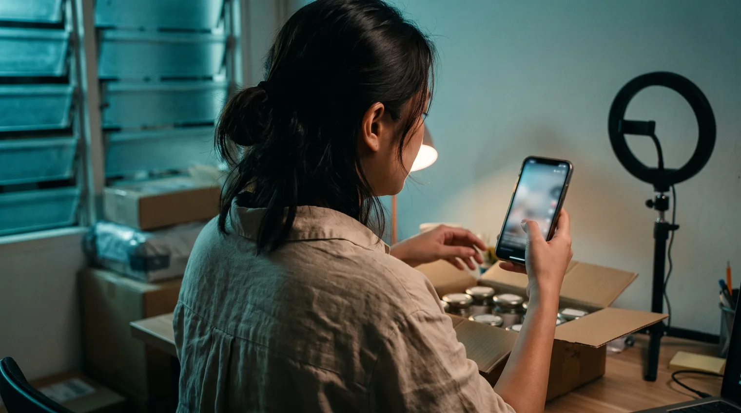 A brand owner at night in a small shoplot office, watching a short video on her phone beside a box of products and an unused ring light